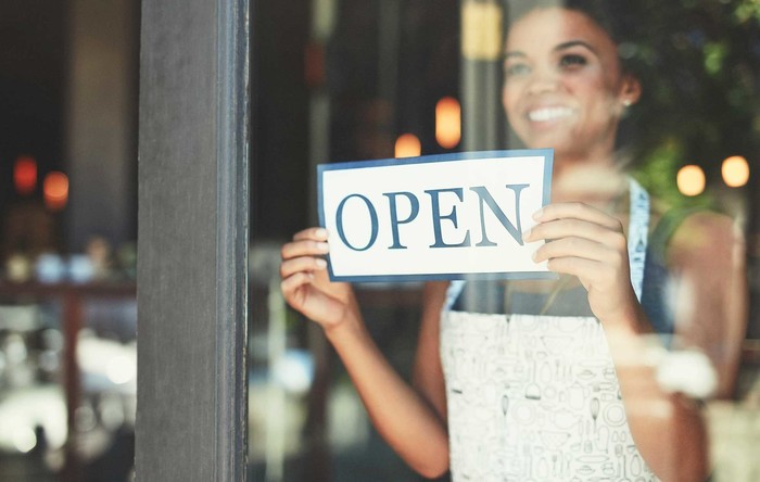 Woman holding sign Open. She bootstrapped her business and it is her first day Open